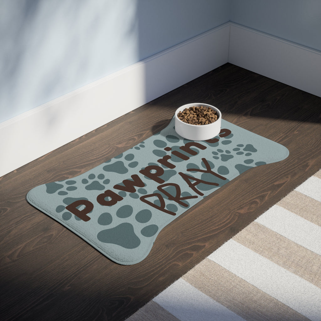 Bone‑shaped plush pet feeding mat with aqua background, dark grey paw print pattern, and brown text “Pawprints Pray”.