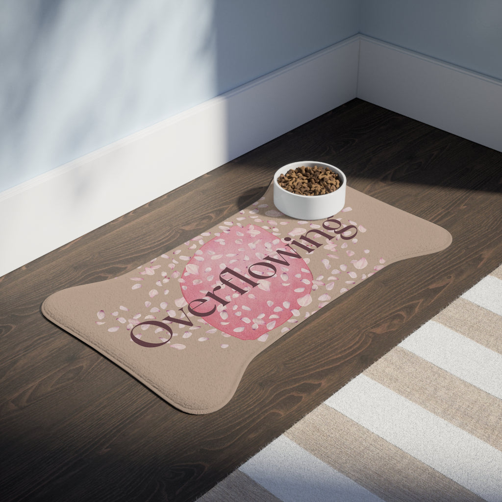 Beige pet feeding mat with pink petal design and the word “Overflowing” printed across the center placed in a room.