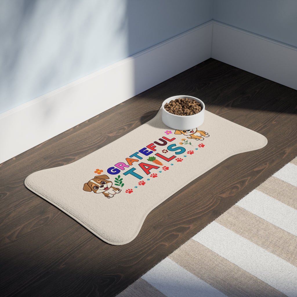 Cream bone-shaped “Grateful Tails” pet feeding mat with colorful design and food bowl placed on dark wood floor next to area rug.