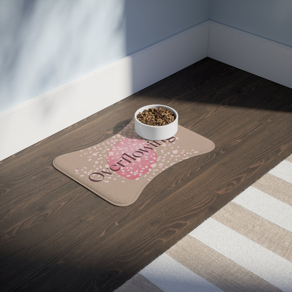 Beige pet feeding mat with pink petal design and the word “Overflowing” printed across the center placed in a room.