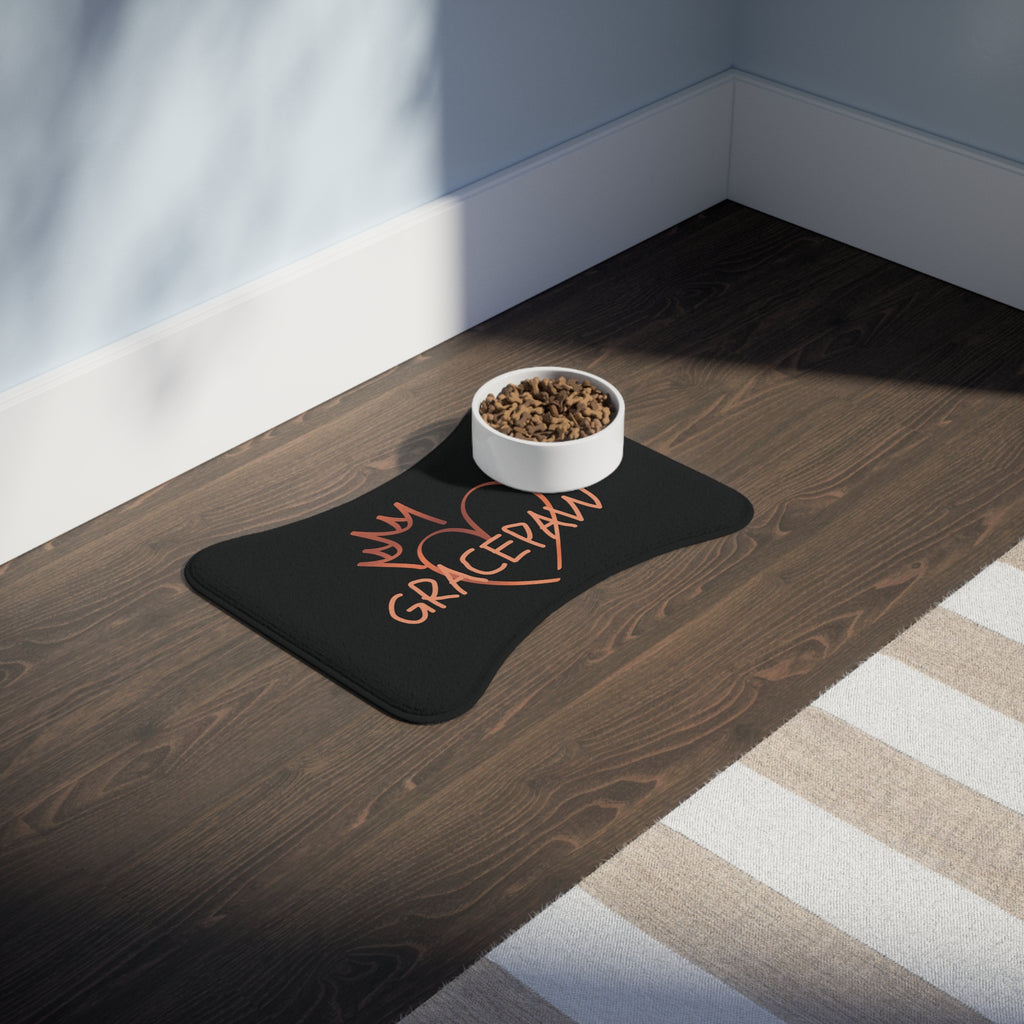 Black bone-shaped pet feeding mat with a heart and crown graphic and the word “GRACEPAW” in peach lettering, designed for comfort and non-slip functionality. The mat is place in a room with a feeding bowl on top. 