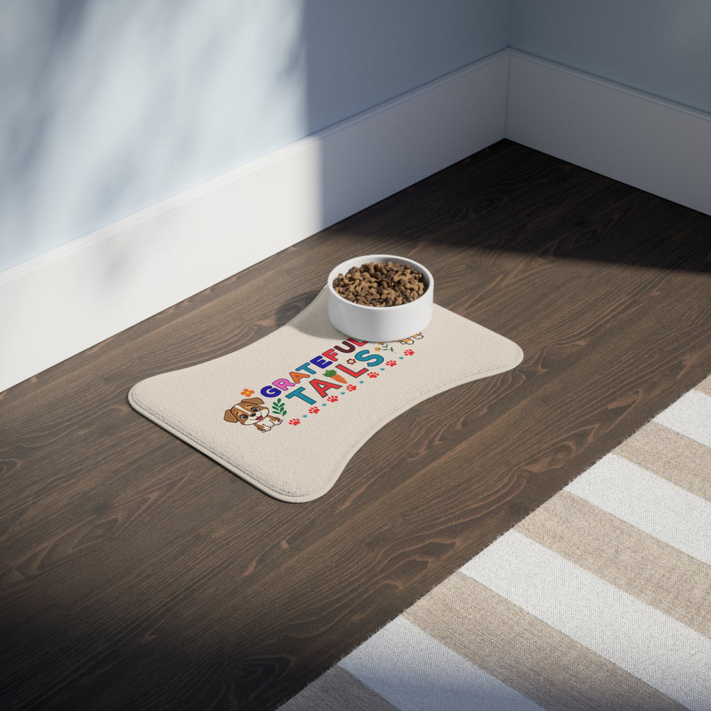 Cream bone-shaped “Grateful Tails” pet feeding mat with colorful design and food bowl placed on dark wood floor next to area rug.