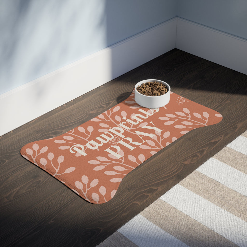 Terracotta bone-shaped pet feeding mat with white floral print and food bowl on dark wood floor near striped rug.