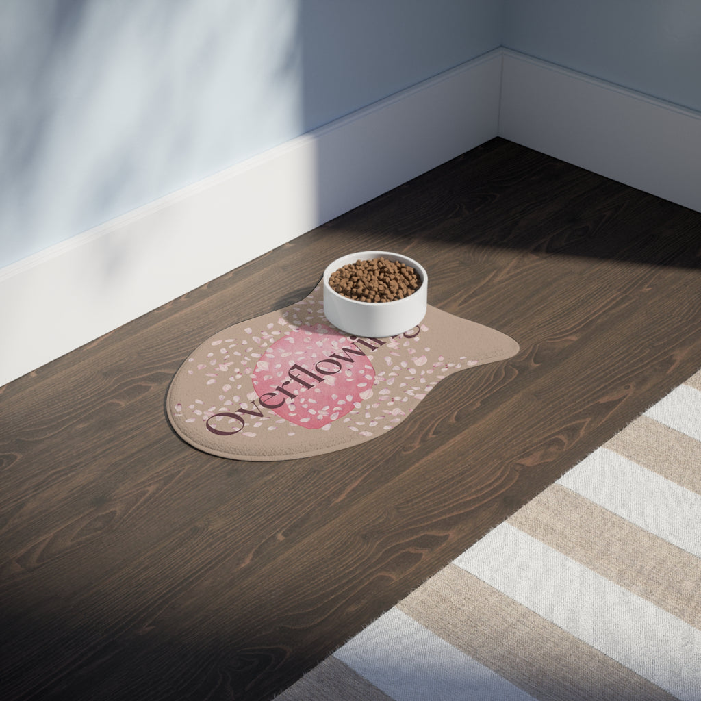 Beige pet feeding mat with pink petal design and the word “Overflowing” printed across the center placed in a room.