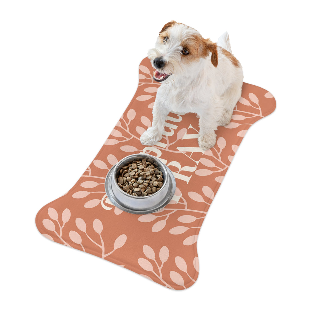 Small white and brown dog sitting beside food bowl on bone-shaped terracotta pet mat with floral design.