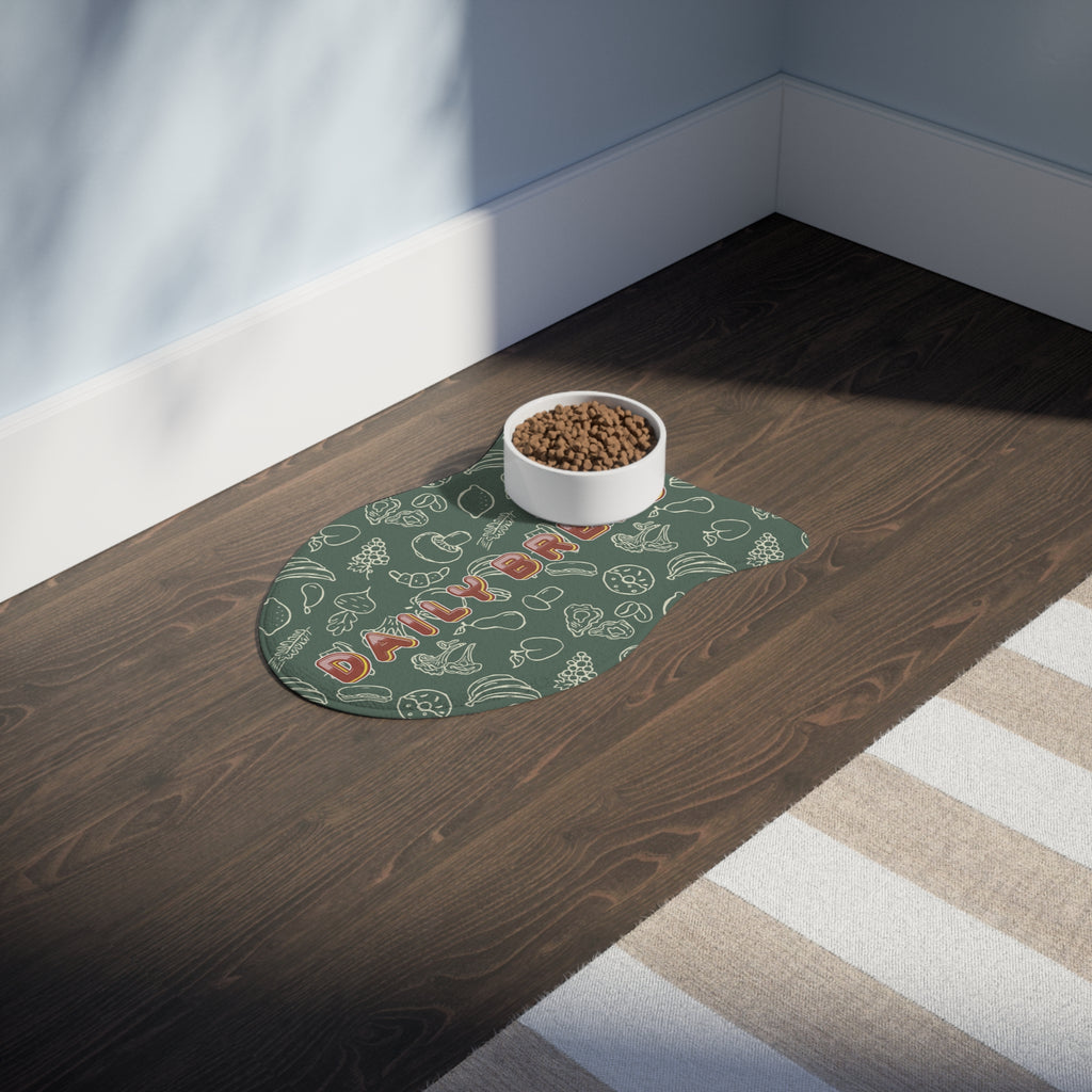 Sage green fish-shaped pet feeding mat with white doodle pattern placed on dark wood floor beside a white food bowl.