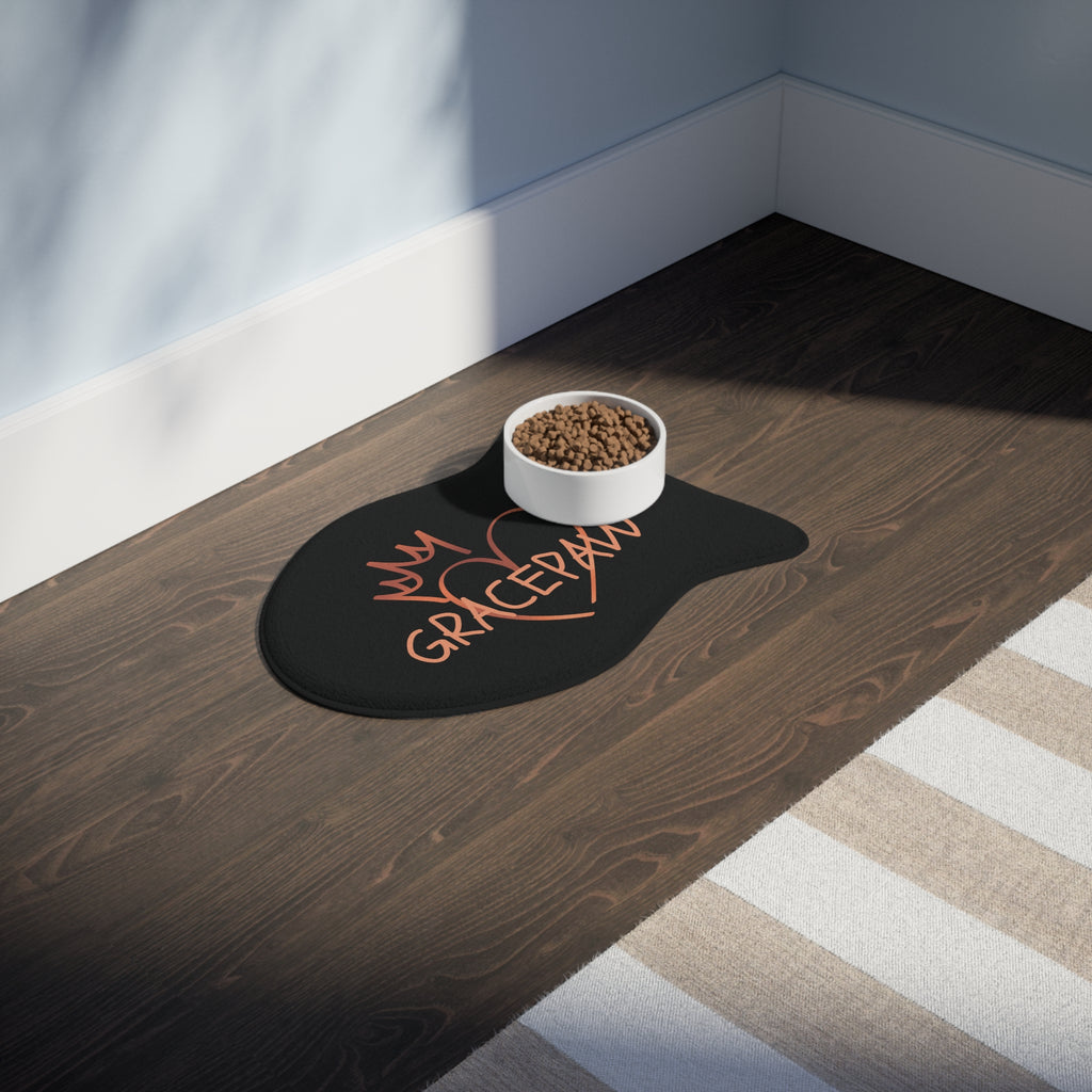Black fish-shaped pet feeding mat with a heart and crown graphic and the word “GRACEPAW” in peach lettering, designed for comfort and non-slip functionality. The mat is place in a room with a feeding bowl on top. 