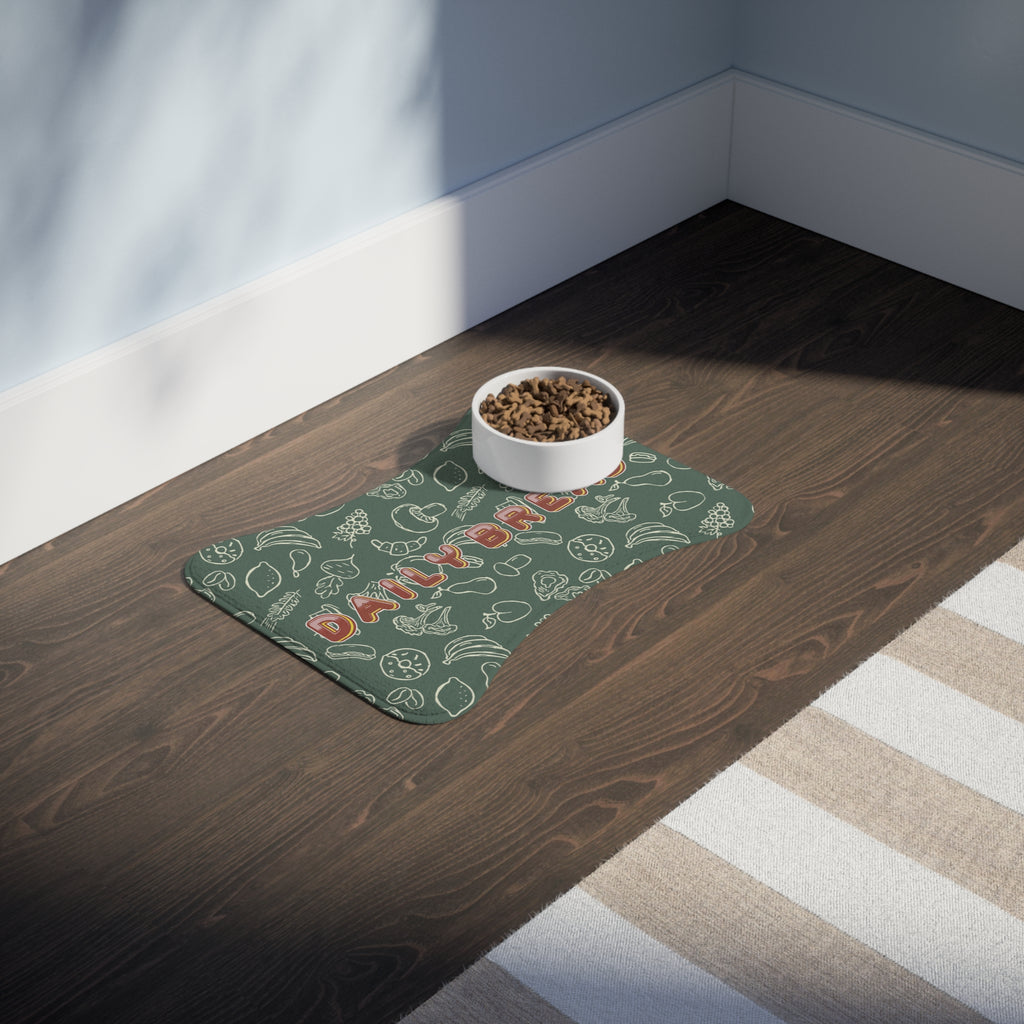 Sage green bone-shaped pet feeding mat with white doodle pattern placed on dark wood floor beside a white food bowl.