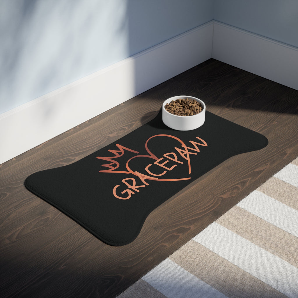 Black bone-shaped pet feeding mat with a heart and crown graphic and the word “GRACEPAW” in peach lettering, designed for comfort and non-slip functionality. The mat is place in a room with a feeding bowl on top. 
