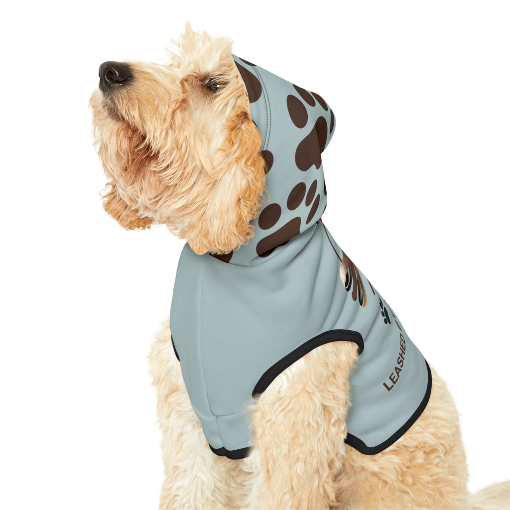 Fluffy light tan dog wearing the blue-gray hoodie, looking up, showing the dark brown paw print pattern on the hoodie and black trim. 