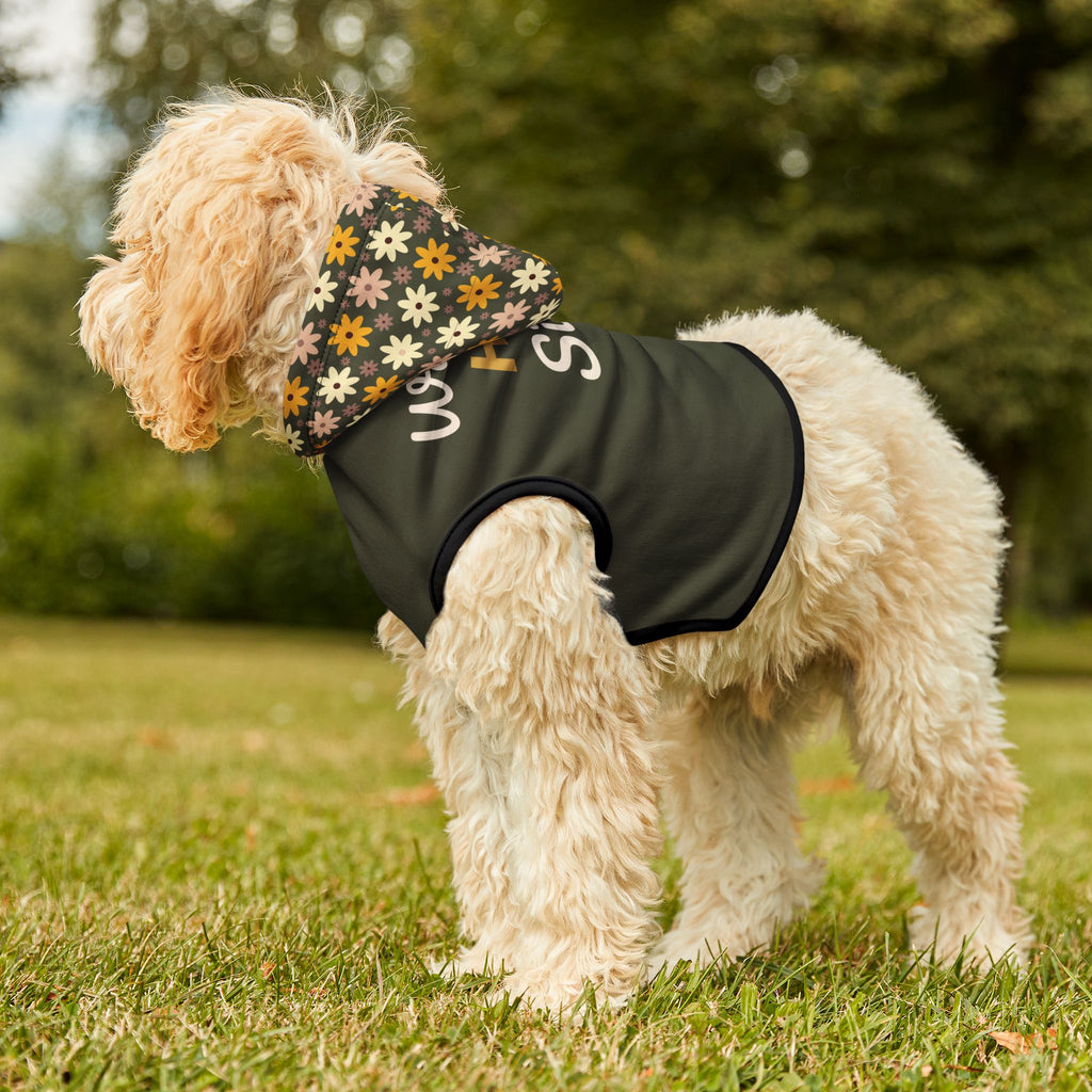 Fluffy light tan dog in a park, wearing an olive green hoodie, looking away, showing the floral design pattern on the hoodie, the graphic, " Walking by HIS Side", and black ribbed trim. 