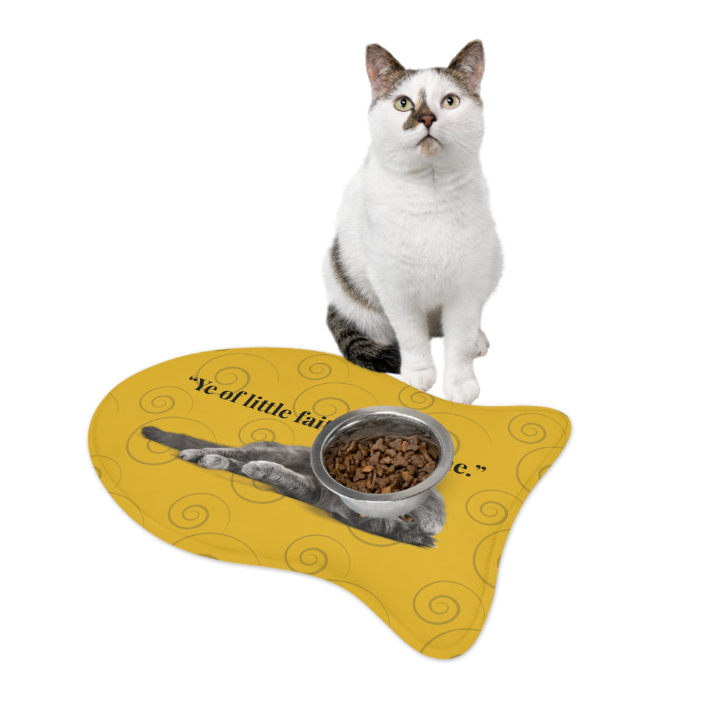 White and gray cat sitting next to yellow fish-shaped feeding mat with humorous faith-based quote and food bowl.
