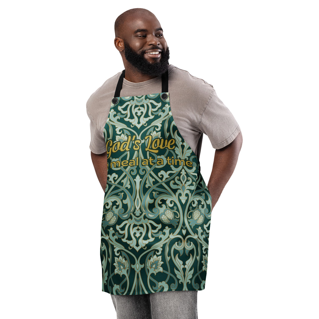 Faith-inspired apron with ornate sage and emerald baroque-style floral pattern, featuring the phrase “God’s Love  One Meal at a Time” in elegant gold lettering across the chest.