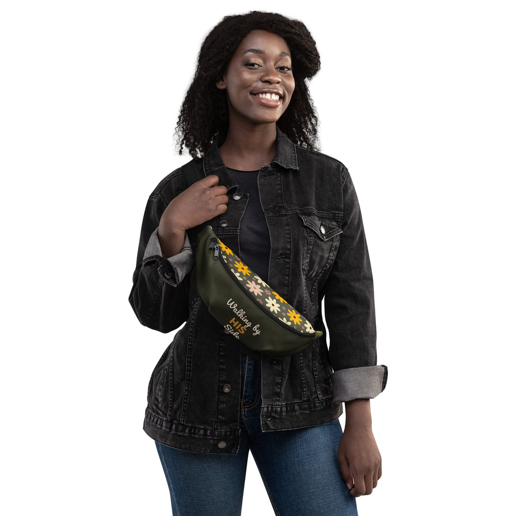 A woman wearing an olive green fanny pack with a floral design and "Walking By HIS Side" across her chest.