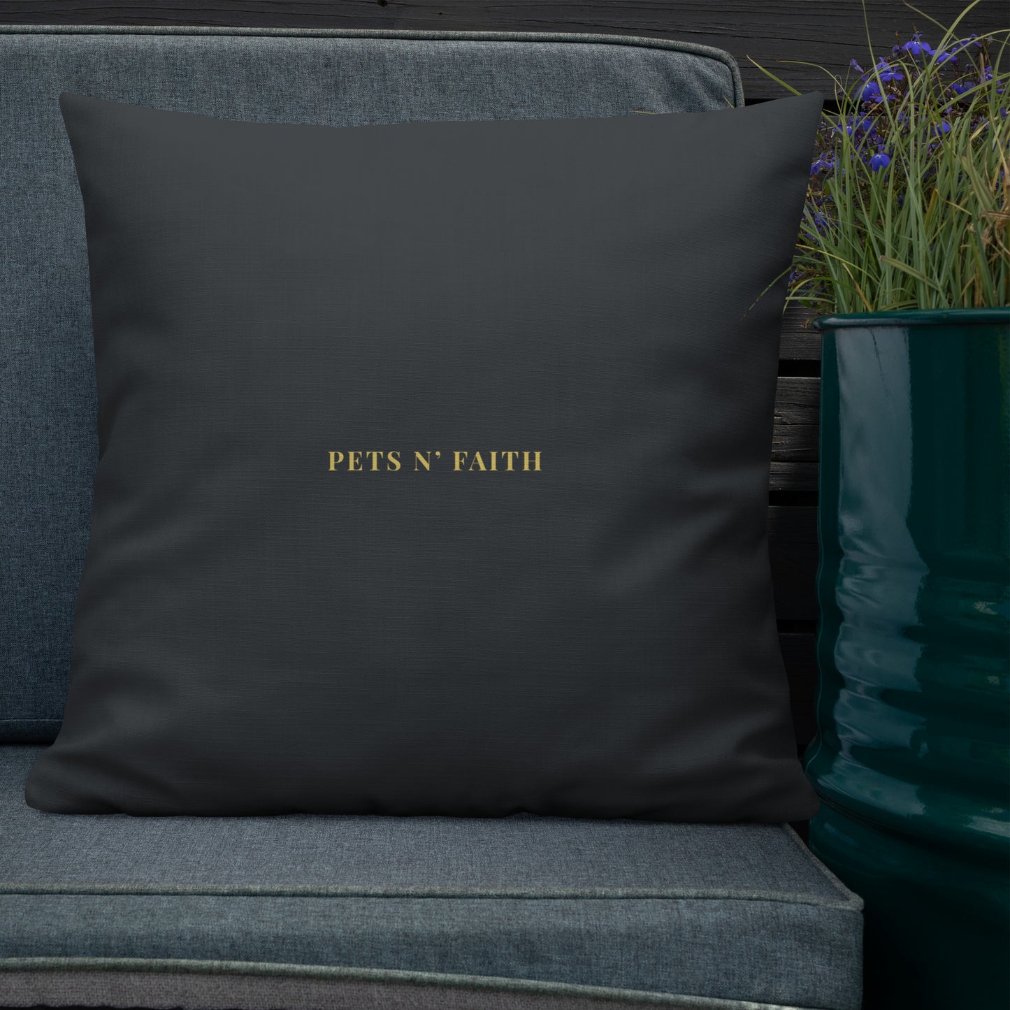 Dark grey pillow with 'PETS N' FAITH' text on a gray couch next to a potted plant.