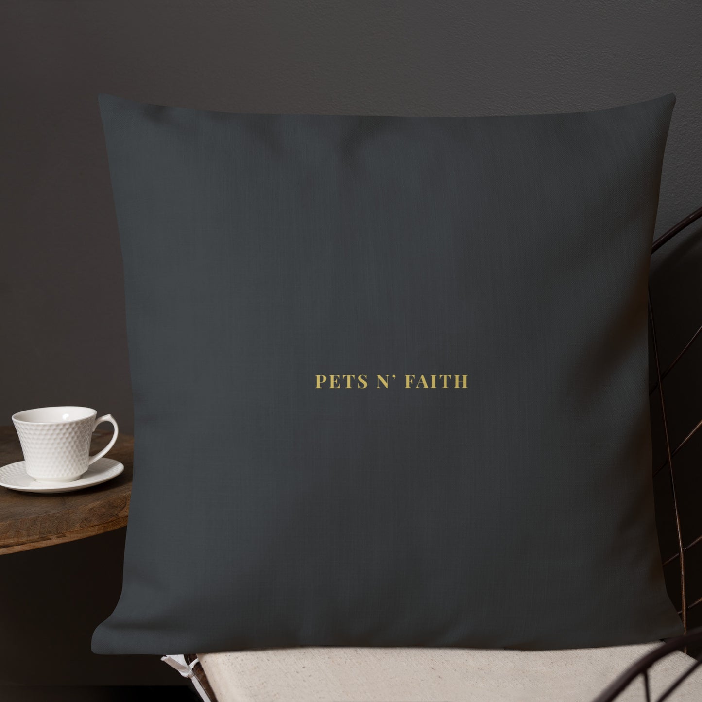 Dark gray pillow with 'PETS N' FAITH' text on a chair with a cup and saucer in the background.