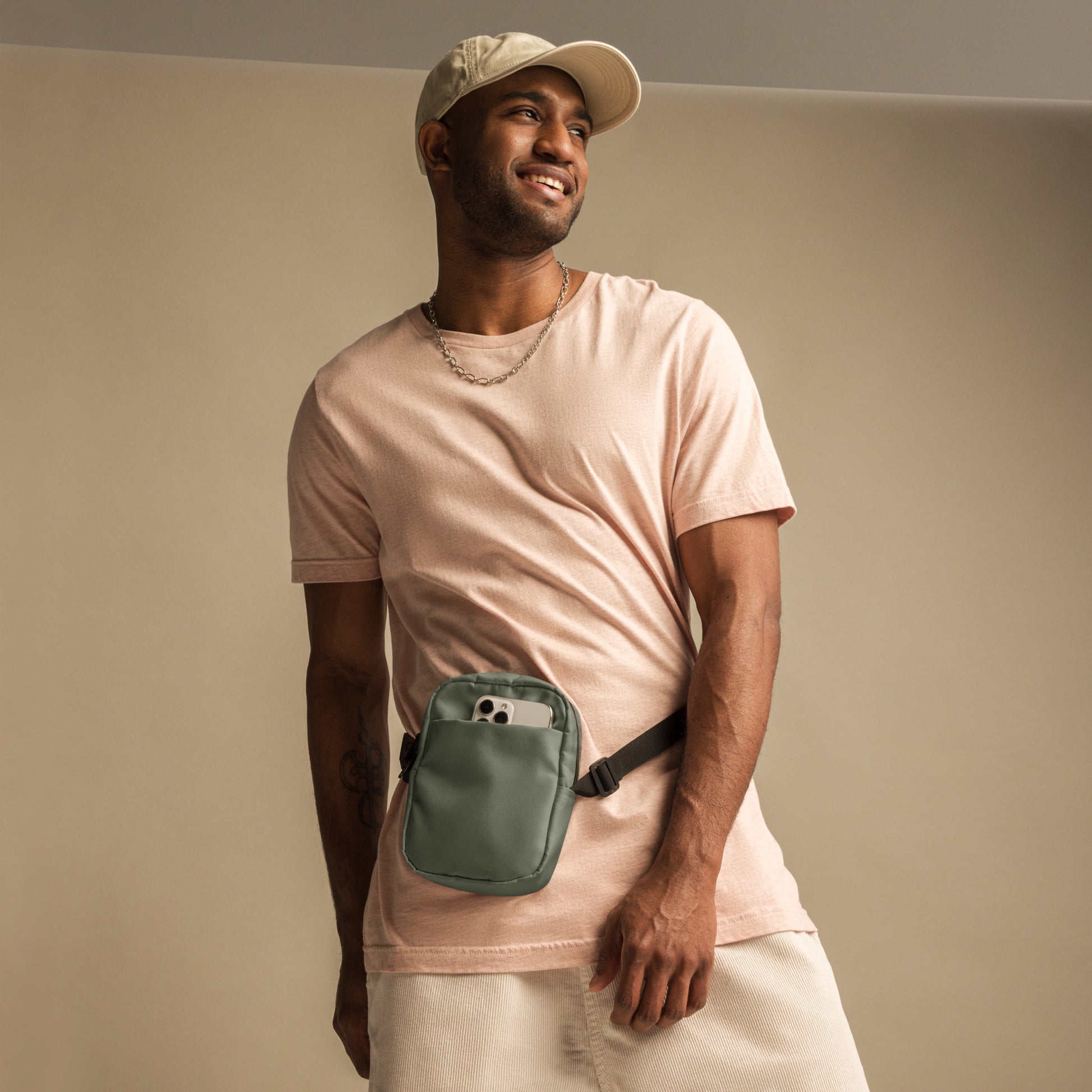 A man wearing a pink t-shirt and beige cap with a military green waist bag on a plain background