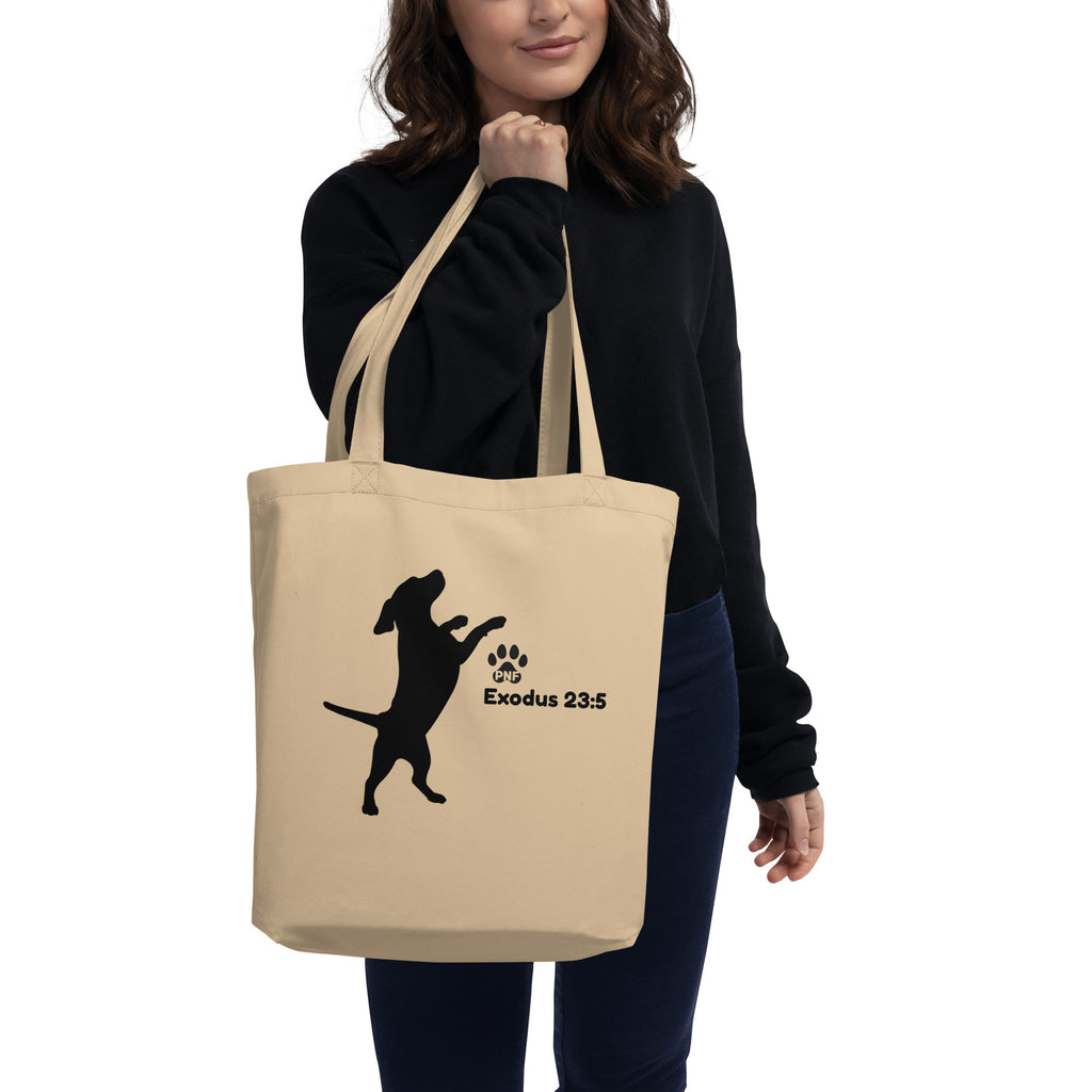 Woman wearing black sweater holding beige tote bag with black dog silhouette and “Exodus 23:5” scripture reference.