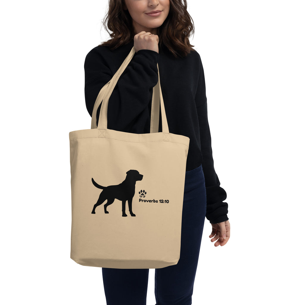 Woman wearing black sweater holding beige tote bag with black dog silhouette and scripture reference "Proverbs 12:10".