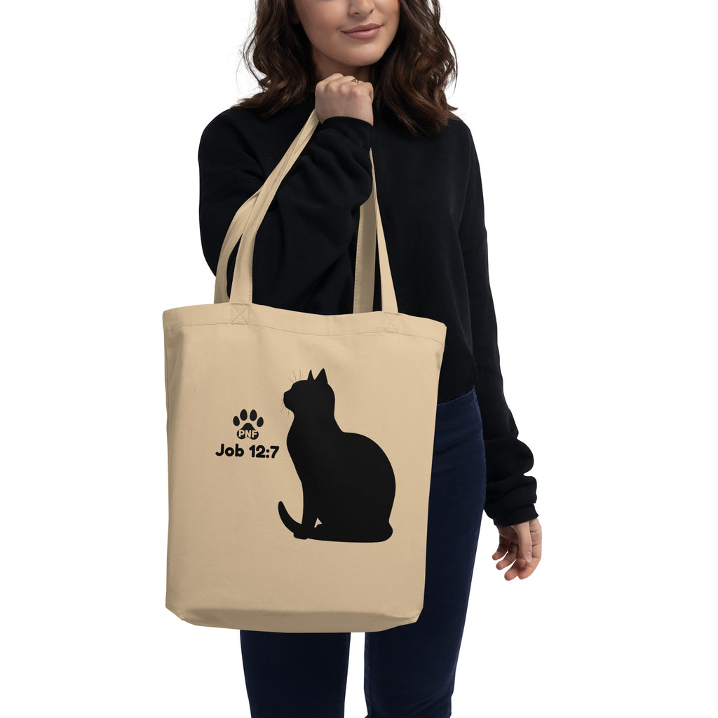Woman holding beige tote bag with black cat silhouette and Job 12:7 scripture design.