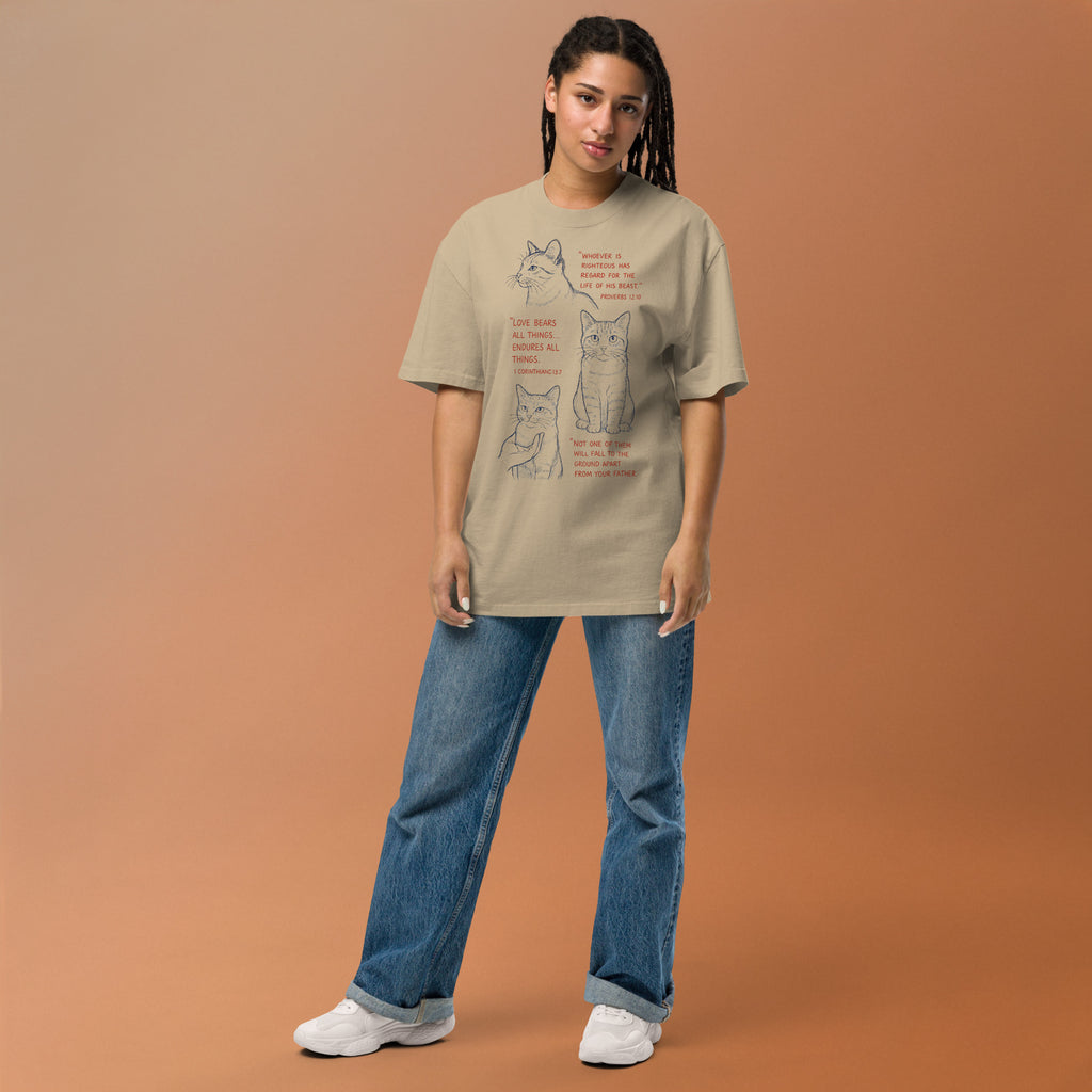 Woman standing with khaki oversized faded t-shirt with cat illustrations and Bible verses, including Proverbs 12:10, 1 Corinthians 13:7, and Matthew 10:29, designed by Pets N Faith.