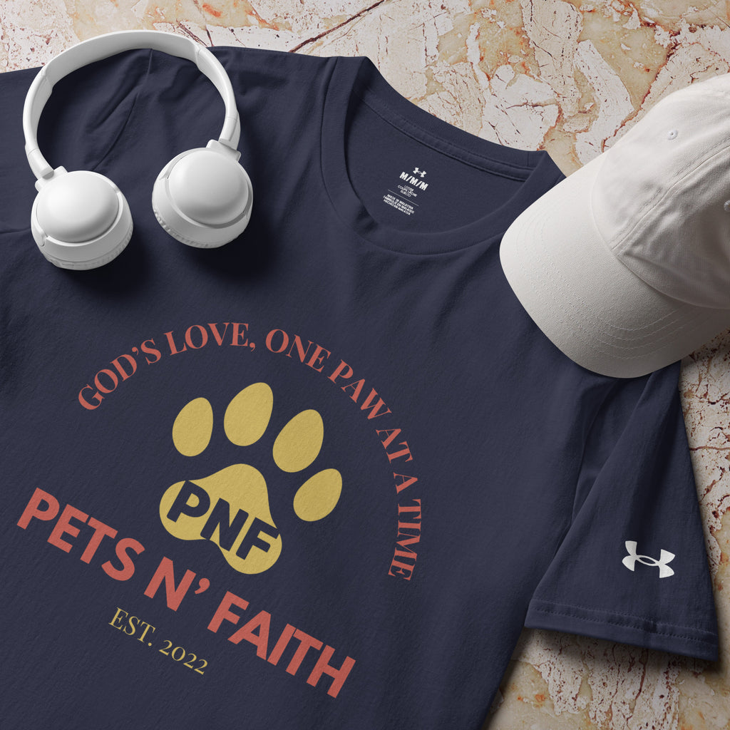 Close-up of navy blue Under Armour Pets N’ Faith Performance T-Shirt with “ God's Love, One Paw at A Time” design, next to a white hat and white headphones. The PNF initials are inside of a gold paw print with the Pets N' Faith  Est. 2022 logo in red text. 