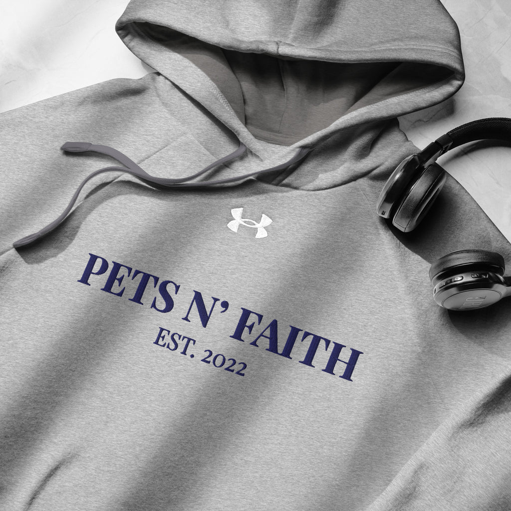 Close-up of light gray Under Armour Pets N’ Faith Hoodie with navy print and drawstring hood, next to wireless headphones