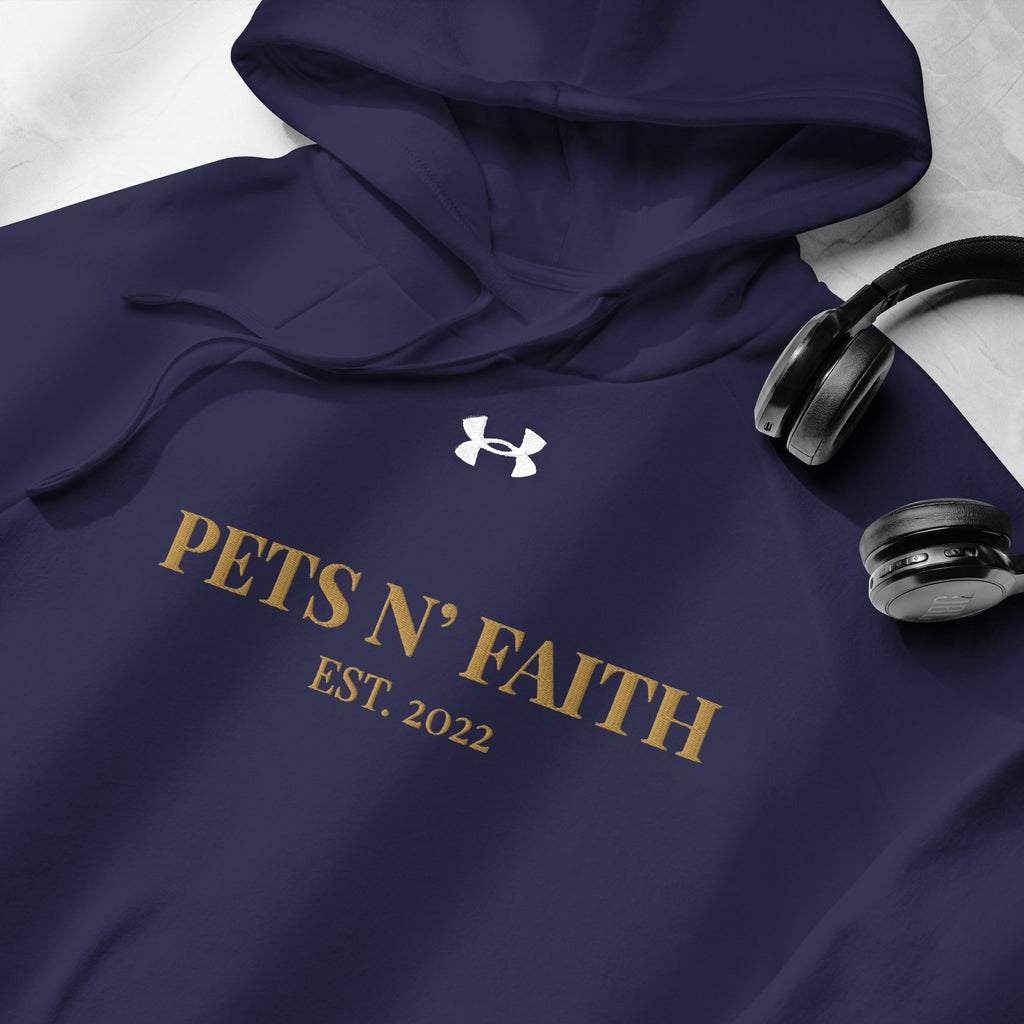 Close-up of  navy blue Under Armour Pets N’ Faith Hoodie with gold print and drawstring hood, next to wireless headphones